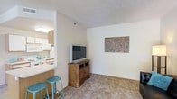 This single-floor residence showcases an open-concept design that seamlessly connects the living area and kitchen. The living space features neutral beige carpeting and light-toned walls, creating a warm and inviting atmosphere. A wooden media console serves as the focal point of the living area, complemented by decorative wall art that adds a subtle touch of character to the space.

The kitchen is partially open to the living area, separated by a breakfast bar with teal counter stools — a functional and stylish dividing element. The kitchen features white cabinetry, white appliances, and light-colored countertops, contributing to a clean and cohesive aesthetic. A ventilation hood is positioned above the stove, and the kitchen benefits from recessed lighting overhead.

The open layout between the two spaces promotes a sense of connectivity and makes the overall footprint feel spacious and practical. The consistent use of light, neutral tones throughout both areas enhances the brightness of the interior. The textured ceiling and visible ventilation duct add functional architectural details to the overall design.