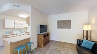 This single-floor residence showcases an open-concept design that seamlessly connects the living area and kitchen. The living space features neutral beige carpeting and light-toned walls, creating a warm and inviting atmosphere. A wooden media console serves as the focal point of the living area, complemented by decorative wall art that adds a subtle touch of character to the space.

The kitchen is partially open to the living area, separated by a breakfast bar with teal counter stools — a functional and stylish dividing element. The kitchen features white cabinetry, white appliances, and light-colored countertops, contributing to a clean and cohesive aesthetic. A ventilation hood is positioned above the stove, and the kitchen benefits from recessed lighting overhead.

The open layout between the two spaces promotes a sense of connectivity and makes the overall footprint feel spacious and practical. The consistent use of light, neutral tones throughout both areas enhances the brightness of the interior. The textured ceiling and visible ventilation duct add functional architectural details to the overall design.