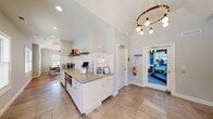 The clubhouse at 9 Covered Bridge Road is a thoughtfully designed single-floor community gathering space that seamlessly blends comfort, functionality, and modern style. The main social area serves as the heart of the clubhouse, anchored by a striking full-height stacked stone accent wall that adds warmth and texture to the open interior. A comfortable sectional sofa sits against the stone wall, creating an inviting lounge space, while vaulted ceilings and light hardwood flooring enhance the airy, spacious feel throughout. The bar and dining area features a natural wood plank ceiling with pendant lighting, a wall-mounted TV, and a mix of communal dining tables and bar-height seating, making it ideal for both casual gatherings and social events. A bright dining nook with large windows allows abundant natural light to fill the space, complementing the warm wood tones of the live-edge dining table. The kitchenette is well-appointed with granite countertops, white subway tile backsplash, open wood shelving, a stainless steel sink, coffee station, and under-counter refrigeration. Glass French doors lead from the lounge area to an outdoor patio, inviting easy indoor-outdoor flow. A dedicated fitness center, visible through a glazed door, rounds out the amenities with cardio and strength training equipment.
