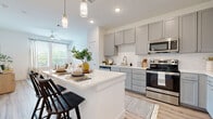 This single-floor residence is thoughtfully designed with a contemporary aesthetic, combining functionality and style across its open layout. The kitchen serves as a central highlight, featuring gray shaker-style cabinetry, a full suite of stainless steel appliances including a refrigerator, range, and over-the-range microwave, and a white quartz countertop island with seating for two. Pendant lighting above the island adds a refined touch, while light wood-look flooring and subway tile backsplash contribute to the clean, modern palette. The kitchen flows seamlessly into the living area, which benefits from large windows that invite abundant natural light and offer views of surrounding greenery. A ceiling fan overhead and generous floor space make this area comfortable and versatile. The bedroom is well-proportioned, featuring a ceiling fan, a window with natural light, and easy access to a dedicated bathroom. The bathroom is organized as a corridor-style space with a gray vanity, white countertop, and direct access to a walk-in closet with built-in shelving and hanging storage. Light wood flooring continues throughout, tying the entire unit together with a cohesive, airy feel.