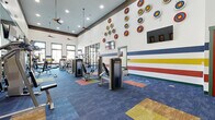 Located at 9500 Silver Collection Cir in Fredericksburg, VA, this well-appointed fitness center offers a comprehensive, single-floor layout designed to support a wide range of workout disciplines. The main workout area is defined by soaring ceilings, ceiling fans, and an abundance of natural light streaming through large, green-trimmed windows that frame views of the surrounding community. Bold horizontal stripes of blue, yellow, and red accent the walls, complemented by a collection of circular target-style wall art that adds a dynamic, energetic character to the space. The floor features a vibrant patchwork of colorful carpet tiles, reinforcing the lively aesthetic. This primary zone is fully equipped with TRUE-brand cardio machines—including treadmills, ellipticals, and stationary bikes—as well as a comprehensive selection of strength training machines, free weights, dumbbells, and medicine balls. A large wall mirror expands the sense of space and supports proper form during exercise. A glass-partitioned interior room provides additional strength training space, while two dedicated side rooms feature durable speckled rubber flooring. One serves as a spin studio with multiple stationary cycling bikes and agility ladder markings, and the other functions as a functional training room with a wall-mounted TV, suspension trainers, and plyo boxes.