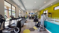 Located at 1502 Partnership Way in Houston, TX, this single-floor fitness center is a comprehensively equipped facility designed to support a full range of workout styles. The space features a bold, contemporary aesthetic, highlighted by vivid chartreuse accent walls, a teal reception counter, and a distinctive hexagonal-patterned carpet in shades of yellow, gray, and taupe that anchors the main floor.

The cardio zone is generously outfitted with treadmills, stationary bikes, rowing machines, and ellipticals. Large floor-to-ceiling windows along the perimeter flood the space with natural light and offer views of the surrounding landscaped grounds, including palm trees and a pool area. Mounted flat-screen televisions keep members entertained during cardio sessions.

The free weights area includes multiple dumbbell racks with a broad weight range, medicine balls, stability balls, and workout benches, accompanied by wall mirrors and high ceilings that enhance the sense of openness. A dedicated functional training zone features a heavy punching bag, a multi-purpose cable and resistance training rig, and a suspension training system, making it suitable for strength and conditioning workouts.

Recessed lighting throughout ensures even illumination across all workout zones. The facility is accessed through glass-panel doors that connect to the outdoor community, reinforcing its open, welcoming character.