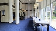 This single-floor space exudes a modern, upscale ambiance defined by its dramatically high ceilings and open layout. Tall white columns anchor the room, contributing to its grand architectural presence. The vivid royal blue carpet creates a bold visual foundation that contrasts elegantly with the bright white furnishings, including sleek dining tables and ghost-style chairs arranged for formal or social gatherings. A long communal white table serves as a central focal point, styled with blue glass vases and floral accents. Sheer floor-to-ceiling drapery panels add a soft, refined texture to the walls, complementing the neutral gray and white color palette. Pendant chandeliers and drum-shade fixtures hang at varying heights, providing layered, atmospheric lighting throughout. A bar or counter area is visible toward the rear of the space, complete with bar-height seating, suggesting amenities suited for entertaining. Large windows along one side allow generous natural light to filter in, enhancing the room's airy and sophisticated feel. The overall design reflects a contemporary aesthetic well-suited for a hospitality, clubhouse, or event venue setting.