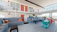 This lively outdoor common area serves as the social heart of a two-story hospitality property, designed with a bold, retro-inspired aesthetic. The spacious deck is finished with gray composite wood planking that runs the length of the courtyard, creating a clean, cohesive foundation throughout the space.

Large built-in bench seating, painted in a cool slate blue, lines the courtyard and is outfitted with striking black-and-white striped cushions accented by colorful throw pillows in red, yellow, and orange. Scattered accent furniture — including vivid teal bistro tables and chairs alongside bold red seating — adds playful pops of color that enliven the entire area.

The surrounding two-story structure frames the deck on multiple sides, with the upper level featuring a white metal balcony railing and a series of signature coral-orange doors that provide strong visual contrast against the white stucco exterior. The lower level mirrors this design, with matching orange doors and louvered shutters opening directly onto the deck.

A white metal staircase connects the ground-level deck to the upper floor, serving as both a functional and architectural element. Open sightlines extend beyond the property's perimeter railing, offering views of the surrounding landscape under open skies. The overall atmosphere is energetic and welcoming, ideal for relaxed outdoor gatherings.