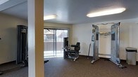 This single-floor fitness space is designed to support a well-rounded exercise routine in a clean, functional environment. The room is outfitted with dedicated strength-training equipment, including a multi-function cable machine and a seated leg exercise unit, offering users a variety of resistance-based workout options. Soft carpet flooring covers the exercise area, providing a comfortable surface underfoot. Natural light enters generously through a large sliding glass door fitted with vertical blinds, offering views of the surrounding neighborhood and creating a bright, open atmosphere. The walls are finished in a neutral tone, complementing the textured ceiling above. Overhead fluorescent lighting supplements the natural light, ensuring the space remains well-lit at all hours. A structural column near the entrance adds an architectural element to the room's layout without impeding functionality. The overall ambiance is practical and welcoming, making it a convenient and accessible fitness facility for everyday use.