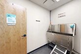 Located at 22 Loso Dr in Cicero, NY, this purpose-built pet wash station is a compact, single-room facility designed specifically for dog grooming and bathing. The room features clean white walls, a drop ceiling with recessed lighting panels, and a durable diamond-plate rubber flooring that provides traction and easy cleanup — ideal for a wet-use environment.

The centerpiece of the space is a large, professional-grade stainless steel Triton dog bath tub, elevated on a sturdy frame and equipped with an integrated access ramp for easy pet entry. The tub features built-in securing straps and a side-mounted water supply with hot and cold controls, color-coded for convenience. An additional wall-mounted faucet and utility sink provide supplementary wash access.

A wall-mounted pet dryer is installed adjacent to built-in white shelving, which offers organized storage for grooming supplies and accessories. A multi-hook leash rack is mounted on the wall near the entrance, providing a practical spot to secure pets before and after bathing. Informational grooming guides and posted station rules are displayed on the walls for easy reference.

The space is accessed through a solid wood door clearly labeled "Dog Wash," connecting it to the broader facility corridor. The overall design is clean, functional, and well-organized, reflecting a professional approach to pet care.