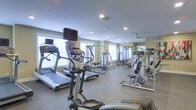 This fitness center is a single-floor facility designed to support a comprehensive workout experience. The space is outfitted with a strong selection of cardio equipment, including treadmills and elliptical machines, each equipped with individual monitor screens for entertainment during workouts. Complementing the cardio section, a range of cable and weight machines provides ample options for strength training. The flooring features a durable, speckled rubber surface well-suited for high-traffic fitness use. Recessed lighting is installed throughout the ceiling, ensuring bright and even illumination across the entire floor. Large windows along the walls invite natural light into the space, contributing to an open and energizing atmosphere. Full-length mirrors are mounted on one wall, a practical feature that allows users to monitor their form during exercises while also enhancing the sense of depth and spaciousness within the room. The neutral wall tones and clean layout give the facility a professional and welcoming appearance. Overall, this fitness center offers a well-organized, functional environment suitable for a wide range of fitness routines and training levels.