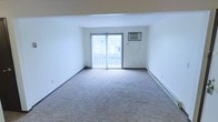 This single-floor space offers a clean and functional layout designed for comfortable everyday living. The main living area is generously proportioned, featuring soft wall-to-wall carpeting that adds warmth underfoot and contributes to a quiet, relaxed atmosphere. Crisp white walls throughout keep the space feeling bright and airy, providing a neutral backdrop well-suited for a variety of interior styles. A prominent sliding glass door serves as the focal point of the living area, allowing abundant natural light to fill the room while providing direct access to a private balcony — an ideal spot for outdoor relaxation. Vertical blinds accompany the sliding door, offering flexible light control and privacy. A wall-mounted air conditioning unit ensures year-round comfort, while a baseboard heating system runs along the perimeter of the room for efficient warmth during cooler months. The overall ambiance is simple and move-in ready, with a straightforward design that prioritizes functionality. The open, uncluttered layout offers flexibility in furniture arrangement, making it easy to personalize the space to suit individual needs. This unit is well-suited for those seeking a practical and comfortable living environment.