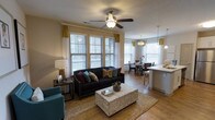 This single-floor residence offers a thoughtfully designed open-concept layout that seamlessly connects the main living areas. The living room serves as the heart of the space, featuring warm hardwood-style flooring, a ceiling fan with light fixture, and an abundance of natural light streaming through large floor-to-ceiling windows dressed with elegant gold and white curtains. The room comfortably accommodates a dark sofa adorned with colorful accent pillows, a teal accent chair, and a mosaic-tiled coffee table anchored by a woven area rug, creating a cozy yet stylish atmosphere. Adjacent to the living area, the dining space offers a welcoming setting for meals, bathed in natural light from additional windows and accented with pendant lighting overhead. The kitchen is positioned just beyond, featuring a breakfast bar with bar seating, pendant lights, and light-colored countertops that add a clean, contemporary touch. Neutral warm-toned walls complement the hardwood-style flooring that flows consistently throughout the space, unifying each area into a cohesive and inviting home environment. The overall ambiance is bright, comfortable, and well-appointed.