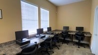 This single-floor computer lab is designed as a functional shared workspace, offering multiple individual workstations arranged along L-shaped, granite-style countertops that line the perimeter walls. Each station is equipped with a desktop computer and monitor, paired with ergonomic rolling chairs that provide comfortable seating for extended use. The room is finished with warm beige walls that contribute to a neutral, focused atmosphere conducive to productivity. Large windows fitted with horizontal blinds allow generous amounts of natural light to fill the space, creating a bright and welcoming environment while still offering control over glare. The flooring features a low-pile patterned carpet that adds a layer of softness underfoot. The layout is efficient, maximizing the available floor space without feeling crowded, and the consistent workstation setup ensures a uniform experience for all users. Overall, the space is well-suited for shared computer access, whether for research, administrative tasks, or general use.