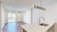 This single-floor space presents a clean, contemporary design characterized by crisp white walls, warm hardwood flooring, and recessed lighting throughout. The open-concept layout connects the kitchen and living areas seamlessly, creating a bright and functional flow. The kitchen serves as a focal point, featuring a striking blue island with a white quartz countertop, an undermount stainless steel sink with a modern pull-down faucet, and a suspended stainless steel range hood overhead. Light wood and dark cabinetry line the walls, complemented by stainless steel appliances including a refrigerator, range, and microwave. The living area is spacious and open, with direct access to a private balcony through sliding glass doors, allowing natural light to fill the room and providing outdoor views. The bedroom spaces are well-proportioned, with large windows that invite abundant daylight and include a ceiling fan for added comfort. A bathroom features grey tile flooring, a white quartz vanity, and a modern rectangular sink with a sleek chrome faucet. Throughout, the design emphasizes clean lines and a neutral palette, creating a cohesive and inviting atmosphere.