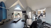 This fitness center is housed within a single-story structure distinguished by a striking vaulted, cathedral-style ceiling that creates an open, airy atmosphere throughout the space. The facility is divided into two distinct zones: a cardio area and a strength training area. The cardio section benefits from large picture windows and a triangular skylight that flood the room with natural light, offering direct views of an outdoor pool and patio area. This section is equipped with multiple treadmills, upright and recumbent stationary bikes, and an elliptical trainer. A wall-mounted flat-screen television provides entertainment during workouts. The strength training area features bold navy blue accent walls, built-in wooden shelving for storage, and a selection of cable machines and weight equipment. Mirrored walls in this zone enhance the sense of space and support proper form during exercise. Throughout both areas, durable dark rubberized flooring provides stability and impact absorption. Track lighting is installed along the ceiling, supplementing the natural light. French doors connect the cardio area to the outdoor space, reinforcing the facility's connection to the exterior amenities. A posted fitness policies sign near the entrance provides facility guidelines for users.