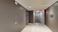 This single-floor space presents a thoughtfully designed interior corridor that combines functionality with a clean, modern aesthetic. The hallway stretches notably in length, creating a sense of depth accentuated by recessed ceiling lighting that illuminates the passage evenly from end to end. A red illuminated exit sign is visible further along the corridor, indicating clear safety compliance and wayfinding measures throughout the space.

The flooring transitions from large-format light tile near the elevator vestibule to a patterned carpet runner along the length of the corridor, adding a layer of visual texture and acoustic comfort. The walls are finished in a warm taupe tone, complemented by crisp white crown molding and baseboards that lend a polished, well-maintained appearance.

An elevator with brushed metal doors is positioned prominently, accompanied by a wall-mounted intercom and call panel, suggesting a multi-unit residential or commercial building. The corridor is lined with evenly spaced doorways on both sides, each framed in white trim for a consistent and orderly look. Overall, the space conveys a clean, professional environment with attention to both safety and design detail.