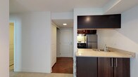 This single-floor space offers a practical and well-organized layout, with the kitchen serving as a central feature. The kitchen is equipped with dark espresso-toned cabinetry that extends from the lower drawers up to overhead storage, creating a cohesive and modern aesthetic. A stainless steel refrigerator and a gas range provide full cooking functionality, while an under-cabinet range hood adds a streamlined, built-in touch. The countertops feature a light speckled surface that contrasts effectively with the darker cabinetry, and a sink with a modern faucet is conveniently integrated into the peninsula-style counter. This counter configuration creates a natural separation between the kitchen and the adjacent living area, which features neutral carpet flooring. A hallway connects the kitchen area to the unit's entry door, which is framed by recessed lighting overhead and hardwood-style flooring underfoot. The clean white walls throughout lend a bright, neutral backdrop, while the overall layout emphasizes efficient use of space and straightforward functionality.