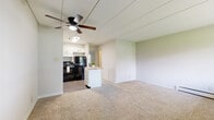 This single-floor unit offers a practical and well-organized layout designed for comfortable everyday living. The open living area is finished with neutral-toned carpet and light green-accented walls, creating a calm and welcoming atmosphere. A ceiling fan with an integrated light fixture provides both airflow and illumination, while panel-style ceiling detailing adds a subtle architectural touch. The space flows seamlessly into the kitchen, which features white cabinetry, dark granite-style countertops, and black appliances including a coil-top electric range and a full-size refrigerator. A compact white kitchen island with a butcher block top provides additional workspace and storage. Wood-style flooring in the kitchen contrasts neatly with the carpeted living area. The bedroom is generously sized with soft carpeting, a large window framing a lush, tree-lined outdoor view, and built-in shelving for added storage. Baseboard heating runs along the perimeter walls, and a wall-mounted air conditioning unit ensures year-round comfort. The bathroom is clean and functional, featuring a single vanity with a granite-patterned countertop, a mirrored medicine cabinet, and a shower-tub combination enclosed by a decorative curtain. Tile-style flooring completes the bathroom's tidy finish.