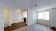 This single-floor residence offers a practical and functional layout with a seamless flow between the main living area and the kitchen. The living room is finished with wall-to-wall carpet flooring, creating a comfortable and inviting atmosphere. A large window allows natural light to fill the space, brightening the neutral white walls and contributing to an open, airy feel. The textured popcorn ceiling adds a classic touch throughout. A ceiling fan with a light fixture is positioned in the transitional area between the living room and kitchen, enhancing both airflow and illumination. The kitchen is accessible through an open archway and features dark cabinetry, black appliances including a dishwasher and range, and a sink positioned along the counter. The kitchen is finished with hardwood-style flooring, contrasting neatly with the carpeted living area. The space benefits from visible HVAC ventilation integrated into the ceiling, ensuring climate comfort. Overall, the layout is clean, efficient, and well-suited for everyday living, with a straightforward design that prioritizes functionality and ease of movement throughout the home.