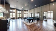 Located at 180 Woodruff Rd in Greenville, SC, this sophisticated amenity and clubhouse space offers a generously proportioned, open-plan layout across a single floor. Dramatically tall ceilings define the character of the space throughout, allowing natural light to pour in through oversized, floor-to-ceiling windows that frame views of the surrounding greenery.

The interior is finished with warm hardwood-style flooring that runs continuously throughout, complemented by a refined palette of muted greens, charcoal tones, and taupe paneled walls. A welcoming lounge area near the entrance features contemporary accent chairs, a lucite side table, and a striking large-scale abstract artwork that sets an upscale tone.

The main gathering area serves as a dynamic game and entertainment hub, anchored by a full-size billiards table and a foosball table, both positioned on a stylish area rug. Multiple mirrored panels along one wall add depth to the space, while tall sheer curtains frame the window walls.

A well-appointed kitchenette area includes a substantial dark-toned island with studded metallic detailing, bar seating for several guests, stainless steel appliances, subway tile backsplash, a wall-mounted television, and a dramatic cluster of decorative pendant lights overhead. An additional lounge nook with an upholstered sofa provides a comfortable retreat. The space is completed by recessed lighting and thoughtfully curated décor throughout.