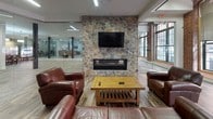 Located at 300 Canal in Lawrence, Massachusetts, this single-floor space masterfully combines the character of a historic mill building with contemporary design and functionality. Original exposed brick walls, tall arched windows, and wide-plank wood-style flooring throughout create a warm industrial aesthetic that pays homage to the building's heritage.

The open layout is anchored by a striking stone accent wall featuring a built-in linear fireplace and wall-mounted television, surrounded by leather seating that forms a comfortable lounge area. Adjacent glass-partitioned spaces offer versatile room options for meetings or private use, maintaining visual connectivity throughout.

A well-appointed kitchen serves the space with white shaker cabinetry, granite countertops, stainless steel appliances including a French door refrigerator, electric range, and microwave. A peninsula with bar-height seating and pendant lighting provides an inviting casual dining or gathering spot.

The dining area accommodates multiple seating arrangements across several tables and chairs, bathed in natural light from the large arched industrial windows that line the brick walls. Decorative touches, including locally themed furnishings, add personality to the space.

Overall, the space presents a cohesive blend of historic charm and modern convenience, making it well-suited for community gatherings, events, or collaborative use.