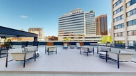 Located on West Baltimore Street in the heart of downtown Baltimore, this well-appointed space offers a compelling combination of modern interior design and striking urban surroundings. The interior is characterized by light hardwood flooring, crisp white walls, and an abundance of natural light delivered through expansive floor-to-ceiling window systems that frame views of surrounding city architecture and an adjacent gravel terrace lined with concrete columns.

The main area is configured to support flexible use, featuring workstation-style desks separated by freestanding privacy panels, upholstered seating in muted tones, and a long communal table suited for collaborative work or meetings. Recessed lighting complements the natural light throughout. A set of double doors with commercial-grade hardware provides clear egress and connects the space to adjoining areas.

A standout feature is access to a spacious rooftop terrace furnished with wicker lounge chairs, chaise lounges, side tables, and bar-height seating arrangements. The terrace offers panoramic views of the Baltimore downtown skyline under open sky, creating an inviting outdoor extension ideal for breaks or informal gatherings. The overall ambiance is clean, contemporary, and well-suited for professional or creative use.