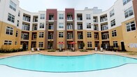 This fitness center is part of a modern, multi-story residential community distinguished by its striking exterior facade, which combines contrasting panels of tan, brown, and deep red siding. The building wraps around a central courtyard anchored by a sparkling, resort-style swimming pool, complemented by poolside seating, umbrella-shaded tables, and lounge chairs — all visible directly from the fitness center's windows.

The fitness center itself is thoughtfully designed to support a comprehensive workout routine. The main exercise floor is spacious and open, finished with durable carpet tile flooring and fitted with recessed lighting and ceiling fans to maintain a comfortable environment during workouts. Equipment includes multiple treadmills, an elliptical, a stationary bike, a rowing machine, cable weight machines, and a dedicated free weight area stocked with a full dumbbell rack and a flat bench.

Large interior windows and pass-through glass panes connect separate zones within the fitness center, creating a sense of openness while allowing natural light to filter through. A wall-mounted flat-screen television adds a modern convenience to the cardio area. An additional windowed alcove overlooks the pool courtyard, providing motivating outdoor views during free weight training. The overall ambiance is clean, bright, and purposefully functional.