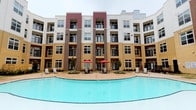 This fitness center is part of a modern, multi-story residential community distinguished by its striking exterior facade, which combines contrasting panels of tan, brown, and deep red siding. The building wraps around a central courtyard anchored by a sparkling, resort-style swimming pool, complemented by poolside seating, umbrella-shaded tables, and lounge chairs — all visible directly from the fitness center's windows.

The fitness center itself is thoughtfully designed to support a comprehensive workout routine. The main exercise floor is spacious and open, finished with durable carpet tile flooring and fitted with recessed lighting and ceiling fans to maintain a comfortable environment during workouts. Equipment includes multiple treadmills, an elliptical, a stationary bike, a rowing machine, cable weight machines, and a dedicated free weight area stocked with a full dumbbell rack and a flat bench.

Large interior windows and pass-through glass panes connect separate zones within the fitness center, creating a sense of openness while allowing natural light to filter through. A wall-mounted flat-screen television adds a modern convenience to the cardio area. An additional windowed alcove overlooks the pool courtyard, providing motivating outdoor views during free weight training. The overall ambiance is clean, bright, and purposefully functional.