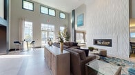 This single-story amenity and leasing space makes a strong architectural impression through its dramatic double-height ceilings and floor-to-ceiling windows that flood the interior with abundant natural light. Large-format tile flooring anchors the main lounge area, where a generously sized sectional sofa and a textured, wave-relief fireplace wall serve as the room's focal point. The sculptural white fireplace surround reaches toward the ceiling, adding a striking design element that complements contemporary artwork displayed throughout.

The lounge transitions seamlessly into a games room featuring dual billiards tables beneath a coffered, warm-lit ceiling and wall-mounted flat-screen televisions. Adjacent to this, a dedicated meeting or presentation room offers a large conference table, bar-height counter seating, and its own display screen—suitable for group gatherings or formal presentations.

A sleek leasing counter, framed by bold navy structural columns and pendant lighting, features a white countertop with bar-height blue accent chairs. Frosted glass doors and glass-walled offices provide a sense of privacy while maintaining visual openness. Large exterior-facing windows and glass entry doors offer views of an outdoor pool area and surrounding landscaping, reinforcing the bright, resort-inspired ambiance throughout.