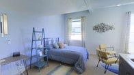 This single-floor bedroom is designed with a soothing color palette, featuring soft blue walls that create a calm and restful ambiance throughout the space. The room centers around a full-sized bed dressed in coordinating blue bedding, accented with decorative pillows in complementary patterns, including a warm geometric print that adds visual interest.

A window positioned along one wall allows natural light to filter in through sheer cream-colored curtains, brightening the space while maintaining a sense of privacy. The window's placement enhances the airy feel of the room, which benefits from a slightly elevated ceiling structure visible in the upper portion of the space.

A teal ladder-style shelving unit stands against one wall, providing both storage and a decorative display area for small décor items. On the opposite side of the room, a yellow accent chair accompanied by a small side table offers a comfortable reading or relaxation nook. A textured wall art piece mounted near the chair adds an organic, sculptural element to the décor.

Neutral carpeted flooring is complemented by a woven area rug beneath the bed, tying the room's elements together cohesively.