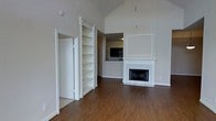 This single-floor residence showcases an open and airy living space anchored by soaring vaulted ceilings that lend a sense of grandeur and volume to the interior. The main living area is centered around a classic white-trimmed fireplace, framed by a decorative paneled surround that adds architectural character and serves as a natural focal point. Rich hardwood flooring flows throughout, providing warmth and continuity across the space.

A built-in bookcase with ornate column detailing stands adjacent to a doorway, offering both functional storage and an elegant design element. The open layout allows for a seamless visual connection between the living area and the kitchen beyond, which is accessible through a pass-through opening. The kitchen features a peninsula counter, providing additional workspace and a casual dining option.

The neutral color palette of soft whites and warm tones throughout the walls and trim creates a clean, versatile backdrop well-suited for a variety of interior styles. The combination of high ceilings, classic architectural detailing, and open sightlines makes this space feel expansive and welcoming, while the hardwood floors and fireplace contribute a sense of comfort and timeless appeal.