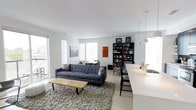 This single-floor space showcases a contemporary, open-plan design that seamlessly blends the living and dining areas into one cohesive environment. The interior is characterized by crisp white walls and ceilings that maximize the sense of light and space, complemented by light-toned hardwood flooring throughout.

The living area centers around a charcoal tufted sofa paired with a slatted wood coffee table, grounded by a plush, textured area rug that adds warmth and contrast to the otherwise minimal palette. Large windows on multiple walls flood the space with natural light, while access to an outdoor area is visible through a sliding glass door, enhancing the connection between interior and exterior.

A tall, dark bookcase serves as a functional focal point, displaying books, framed artwork, and decorative objects, adding depth and character to the space. The dining zone features pendant lighting suspended from the ceiling, casting focused light over a white countertop or island that anchors the area. Throughout, the design maintains a consistent mid-century modern aesthetic, balancing clean geometry with carefully curated furnishings for a polished yet livable atmosphere.