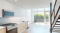 This contemporary two-story residence is thoughtfully designed with a clean, minimalist aesthetic throughout. The ground floor opens into a bright, open-plan living and kitchen area, where floor-to-ceiling glass panels flood the space with natural light and offer direct views of lush greenery beyond a private outdoor area. The kitchen is outfitted with modern flat-front cabinetry in white and warm wood tones, a white quartz countertop, stainless steel appliances including a gas range and dishwasher, and a sleek undermount sink. Light hardwood flooring runs throughout the main living areas, complementing the all-white walls and recessed lighting overhead.

A striking open-tread staircase with a steel frame and cable railing connects the two floors, serving as both a functional element and an architectural focal point. The upper level features an open loft-style bedroom or living space with exposed beams, hardwood floors, and an open railing overlooking the floor below. A well-organized walk-in closet with built-in shelving and hanging rods provides ample storage. The two bathrooms are finished with neutral tile, floating wood-toned vanities, backlit mirrors, and modern fixtures, with one featuring a frameless glass walk-in shower. A hallway connects the upper-level rooms efficiently, completing this well-appointed multi-level residence.