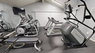 Located at 1401 S Harbor Blvd in La Habra, California, this fitness center offers a well-equipped exercise space designed to support a well-rounded workout routine. The room features a vaulted ceiling that adds a sense of openness, with clean gray walls and durable rubber flooring throughout, creating a functional and practical environment.

The cardio section is outfitted with multiple Matrix-brand treadmills and elliptical trainers, providing a variety of aerobic options for users. Vertical blind-covered windows line one wall, offering light control while maintaining privacy. Overhead linear LED lighting ensures the space is brightly and evenly lit at all times.

Strength training is supported by a multi-cable functional trainer station, a flat weight bench, and a selection of free weights and attachments. Spin/cycling bikes are also available for low-impact cardiovascular workouts. A large wall-spanning mirror runs along one side of the room, visually expanding the space and allowing users to monitor their form during exercise.

A mounted flat-screen television is positioned for easy viewing from the cardio equipment area. Fitness rules and emergency contact information are posted on the wall, reflecting a commitment to user safety. The space is efficiently arranged to maximize equipment variety and usability within its compact footprint.