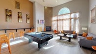 This single-floor space combines entertainment and relaxation in a thoughtfully designed open layout. The room immediately draws attention with its dramatically high ceilings, which amplify the sense of openness and grandeur throughout. A striking mosaic tile column anchors the space, housing a built-in fireplace and a wall-mounted television, serving as a bold focal point that blends function with design.

A full-size pool table in dark tones occupies a central position, making it a natural hub for recreation. Adjacent to it, a casual bar-height shelf with orange stools provides a convenient spot for spectators. The lounge area features a plush sofa, an accent chair in a warm orange hue, and a round area rug atop light wood-toned flooring, creating an inviting conversation zone.

Natural light pours in generously through a large arched window dressed with sheer curtains, softening the brightness while framing outdoor greenery. The warm earth-toned walls and carefully curated artwork contribute to a cohesive, welcoming ambiance that balances lively entertainment with comfortable leisure.