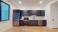 This single-floor unit offers a thoughtfully designed layout with contemporary finishes throughout. The kitchen serves as a central highlight, featuring a full suite of stainless steel appliances, including a French door refrigerator, electric range with over-the-range microwave, and dishwasher. Dark charcoal shaker-style cabinetry extends from floor to ceiling, providing generous storage, while crisp white countertops create a striking contrast. A matte black faucet at the single-basin sink adds a refined touch, and a tile backsplash completes the kitchen's polished aesthetic. Light hardwood flooring runs throughout the main living areas, contributing warmth and continuity to the space. Recessed lighting illuminates the open layout, which includes an expansive main room with exposed ductwork as an industrial design accent, white walls, and multiple interior doors leading to additional areas. A window allows natural light into the kitchen. The bathroom features a clean, minimalist design with white subway tile surrounding a full tub-shower combination, matte black fixtures and hardware, and hexagonal gray tile flooring, creating a cohesive and modern feel throughout the unit.