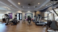This fitness center at 8108 Vinoy Blvd in Charlotte, NC offers a thoughtfully designed single-floor layout that accommodates a wide range of workout styles. The space is organized into distinct exercise zones connected by elegant arched doorways, giving the facility a polished, upscale feel. Tray ceilings with crown molding, recessed lighting, and a ceiling fan contribute to an open and comfortable atmosphere throughout.

The main cardio area houses multiple treadmills, elliptical trainers, and a stationary bike, all positioned to take advantage of natural light from large windows. A wall-mounted flat-screen TV provides entertainment during workouts. The free weights section features a full rack of dumbbells in varying sizes, complemented by stability balls and a dedicated floor mat area for stretching or functional training. Weight benches and a cable machine offer additional strength-training options.

Floor-to-ceiling mirrors in the free weights zone enhance the sense of space and allow users to monitor their form. The facility features wood-look flooring throughout, adding warmth to the functional environment. Entry is provided through sleek double glass doors, offering views of the surrounding outdoor area. Medicine balls on a dedicated rack near the entrance round out the comprehensive equipment offerings, making this fitness center a well-appointed resource for a full-body workout.