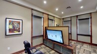 This single-floor entertainment lounge offers a thoughtfully designed space centered around a large flat-screen television mounted on a rich, dark wood media console with frosted glass cabinet doors below. The room's most distinctive architectural feature is its custom wood-framed wall paneling system, which incorporates roller shades and frosted glass panels, creating a structured, refined aesthetic throughout the space. Recessed lighting illuminates the room evenly from a tray ceiling, lending a warm and polished ambiance. The flooring consists of a patterned, multi-toned carpet tile that adds visual texture and complements the warm wood tones of the trim and built-in furnishings. Curated framed artwork adorns the walls, including a vibrant comic-style print and a bold abstract piece, contributing to the room's creative character. An ergonomic gaming chair provides comfortable seating positioned toward the screen, making the space well-suited for interactive entertainment. A glazed wood-framed door connects this lounge to an adjacent dining or meeting area, visible through the glass, suggesting this space is part of a larger multi-use facility. The overall design balances functionality with a refined, contemporary aesthetic.