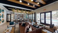 Located at 3529 Adirondack Way in Wilmington, NC, this single-floor community clubhouse is a thoughtfully designed amenity space that blends modern aesthetics with warm, inviting elements. The main lounge anchors the space with a tufted leather sofa, velvet accent chairs, and natural wood stump side tables arranged atop layered area rugs. Striking black-painted exposed beam ceilings contrast beautifully with warm wood tones and globe pendant lighting, creating a sophisticated yet comfortable atmosphere. Large French doors and floor-to-ceiling windows flood the lounge with natural light and provide views to the outdoor surroundings. A built-in bar area features high-top seating alongside open shelving adorned with trailing greenery and curated décor. A café-style dining zone showcases a teal upholstered banquette, marble-top bistro tables, black iron café chairs, and a full wall of white built-in shelving with a mounted TV. A secondary lounge and coworking area features bold emerald green accent walls, exposed wood beam ceilings, woven pendant lighting, swivel lounge chairs, and floating wood shelves with styled vignettes. A dedicated workspace with a built-in desk provides functional seating for multiple users. Cohesive teal, natural wood, and cream tones unify the entire space, reflecting a curated coastal-contemporary design sensibility.