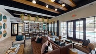 Located at 3529 Adirondack Way in Wilmington, NC, this single-floor community clubhouse is a thoughtfully designed amenity space that blends modern aesthetics with warm, inviting elements. The main lounge anchors the space with a tufted leather sofa, velvet accent chairs, and natural wood stump side tables arranged atop layered area rugs. Striking black-painted exposed beam ceilings contrast beautifully with warm wood tones and globe pendant lighting, creating a sophisticated yet comfortable atmosphere. Large French doors and floor-to-ceiling windows flood the lounge with natural light and provide views to the outdoor surroundings. A built-in bar area features high-top seating alongside open shelving adorned with trailing greenery and curated décor. A café-style dining zone showcases a teal upholstered banquette, marble-top bistro tables, black iron café chairs, and a full wall of white built-in shelving with a mounted TV. A secondary lounge and coworking area features bold emerald green accent walls, exposed wood beam ceilings, woven pendant lighting, swivel lounge chairs, and floating wood shelves with styled vignettes. A dedicated workspace with a built-in desk provides functional seating for multiple users. Cohesive teal, natural wood, and cream tones unify the entire space, reflecting a curated coastal-contemporary design sensibility.