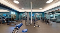 Located on S Alton St in Denver, Colorado, this fitness facility is designed to support a comprehensive workout experience across a single, expansive floor. The main exercise area is generously proportioned, featuring durable rubber flooring and a drop ceiling with recessed circular light fixtures that provide consistent, even illumination throughout the space. Teal-painted accent walls paired with floor-to-ceiling mirrors visually expand the room while allowing users to monitor their form during exercise. High clerestory windows along the upper walls introduce natural light and offer views of the surrounding tree canopy.

The strength training zone is extensively equipped with Hoist-brand cable machines, multi-functional cable stations, adjustable benches, and a full rack of dumbbells, accommodating a wide range of resistance training routines. Structural columns are integrated throughout the open floor plan, defining workout zones without interrupting the overall flow of the space.

A glass-walled studio area separates a dedicated movement or yoga space from the main floor, featuring patterned carpet, floor cushions, and a striking decorative climbing wall with colorful holds that serves as a prominent visual focal point. This transparent partition maintains an open, connected feel while providing acoustic separation for classes or individual sessions.