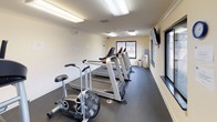 This single-floor fitness center is designed to support a comprehensive workout routine within a clean, efficiently organized space. The interior features neutral cream-colored walls accented by white crown molding and baseboard trim, giving the facility a polished appearance. Durable dark rubber flooring covers the main exercise areas, providing both safety and functionality underfoot.

The cardio zone is well-appointed with multiple treadmills, stationary bikes, and an elliptical machine, all positioned to take advantage of the natural light streaming through several large sliding windows. These windows frame views of a landscaped outdoor area with mature trees, adding a pleasant visual element during workouts. A wall-mounted television is also present for entertainment.

The strength training area includes a multi-station cable machine, a weight bench, and additional resistance equipment, offering a variety of exercise options. Overhead fluorescent lighting ensures the entire space is evenly and adequately illuminated throughout.

A dedicated entryway with a glass-panel door, carpet flooring, and clearly marked exit signage provides a welcoming and safe transition into the facility. A small desk and informational boards are also present, offering a practical reference area for users.