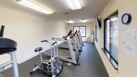 This single-floor fitness center is designed to support a comprehensive workout routine within a clean, efficiently organized space. The interior features neutral cream-colored walls accented by white crown molding and baseboard trim, giving the facility a polished appearance. Durable dark rubber flooring covers the main exercise areas, providing both safety and functionality underfoot.

The cardio zone is well-appointed with multiple treadmills, stationary bikes, and an elliptical machine, all positioned to take advantage of the natural light streaming through several large sliding windows. These windows frame views of a landscaped outdoor area with mature trees, adding a pleasant visual element during workouts. A wall-mounted television is also present for entertainment.

The strength training area includes a multi-station cable machine, a weight bench, and additional resistance equipment, offering a variety of exercise options. Overhead fluorescent lighting ensures the entire space is evenly and adequately illuminated throughout.

A dedicated entryway with a glass-panel door, carpet flooring, and clearly marked exit signage provides a welcoming and safe transition into the facility. A small desk and informational boards are also present, offering a practical reference area for users.