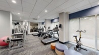 This fitness center offers a comprehensive workout environment designed to accommodate a wide range of exercise routines. The space is laid out across a single open floor, providing ample room for both cardio and strength training activities. A row of treadmills lines one side of the room, accompanied by additional cardio equipment including stationary cycling bikes. On the opposite end, a full rack of dumbbells and a multi-station cable machine cater to free weight and resistance training needs, while a wall-mounted mirror enhances the sense of space and allows users to monitor their form. A dedicated open area with minimal equipment provides flexible space for stretching, yoga, or floor-based exercises. The room features neutral gray and white tones accented by navy blue wall panels, with patterned carpet flooring throughout. Recessed lighting in the drop-panel ceiling provides bright, even illumination across the entire area. Large windows allow natural light to filter in, creating an energizing atmosphere. A motivational fitness-themed mural decorates one wall, contributing to the dynamic ambiance of the space. A built-in water fountain and convenient seating near the entrance round out the practical amenities of this well-appointed facility.