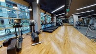 This fitness center is a thoughtfully designed, single-level facility that balances modern aesthetics with comprehensive workout functionality. The space is defined by its warm hardwood flooring, which runs throughout the cardio zone and transitions to a carpeted surface in the dedicated strength training area, clearly delineating each section.

The cardio zone is generously proportioned, lined with a row of Life Fitness treadmills, elliptical cross-trainers, stair climbers, and stationary and spin bikes, all positioned along expansive floor-to-ceiling windows. These windows flood the space with natural light and offer views of lush greenery outside, creating an inviting atmosphere. Wood-paneled accent walls add warmth and texture throughout.

The strength training area is equipped with multiple selectorized cable and weight machines and features large wall-mounted mirrors that visually expand the space. A rowing machine and open floor space for functional training are also present. A mounted flat-screen television and ceiling fan are noted in this zone.

Overhead, a dark exposed ceiling with linear LED lighting strips and pendant fixtures provides a contemporary industrial feel. The entrance area features a brick surround and glass double doors, adding architectural character to this well-appointed facility.