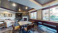 This well-appointed single-floor communal lounge is designed with both comfort and entertainment in mind. The main gathering area features a tray ceiling with dark, contrasting paint accents and crown molding, lending a sophisticated architectural detail overhead. A central fireplace with a mosaic tile surround and built-in cabinetry serves as the focal point of the room, flanked by large windows that invite natural light and offer views of the surrounding exterior. Patterned carpet in cool blue and gray tones adds texture and visual depth throughout the space.

Seating options are varied and plentiful, including leather lounge chairs, a plush sofa with accent pillows, and multiple dining tables with industrial-style chairs — all suited for casual gatherings or organized events. A full-length shuffleboard table runs along one windowed wall, providing a dedicated recreation zone.

The kitchenette area is fully equipped with rich dark wood cabinetry, granite countertops, a deep blue glass tile backsplash, and stainless steel appliances including a refrigerator and wall oven. A bar-height island with pendant lighting and bar stools creates an additional social hub. Decorative wall art and thoughtfully placed accent lighting complete the polished, contemporary aesthetic of this versatile amenity space.
