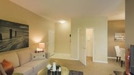 This single-floor residence offers a warm and inviting atmosphere defined by its neutral color palette and soft, wall-to-wall carpeting throughout. The living area serves as the central hub of the space, furnished with a comfortable sofa accented by decorative pillows and complemented by a low coffee table that anchors the seating arrangement. A table lamp provides soft, ambient lighting that enhances the room's cozy feel alongside a flush-mounted ceiling light.

The layout features multiple doorways that connect to additional areas of the residence. One doorway leads to what appears to be an entryway or foyer, distinguished by tiled flooring that contrasts with the carpeted living area, providing a practical transition zone. A second open doorway reveals a connecting room, offering a sense of flow and accessibility throughout the space.

The walls are finished in a soft, warm beige tone that contributes to the overall welcoming ambiance. White-painted door frames and six-panel doors add a clean, classic architectural detail throughout. The space is efficiently organized, making practical use of its layout while maintaining a comfortable and livable feel suited for everyday living.