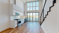 Located at 901 N Vine St in the heart of Hollywood, Los Angeles, this contemporary two-story loft offers a striking open-concept layout defined by soaring double-height ceilings and an abundance of natural light. The main level centers around a seamless living and kitchen area, where large floor-to-ceiling windows and glass sliding doors flood the space with daylight and frame views of the surrounding cityscape and hillside. The sleek, modern kitchen is outfitted with white flat-front cabinetry, white quartz countertops, a subway tile backsplash, and a full suite of stainless steel appliances including a gas range, built-in microwave, dishwasher, and refrigerator. Warm-toned hardwood flooring runs throughout the main level, complementing the clean, minimalist aesthetic. A dramatic staircase with a bold black metal frame and cable railing connects the two floors, serving as a design focal point. The lower level also includes a clean, modern bathroom featuring a floating white vanity, integrated sink, and large mirror. Upstairs, the loft-level space is carpeted and open to below via a half-wall railing, offering flexibility as a bedroom or lounge area with recessed lighting and windows that continue to invite natural light throughout.