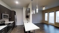 Located in Dallas, this modern single-floor space showcases a thoughtfully designed open-concept layout that seamlessly blends style and functionality. The kitchen is a standout feature, boasting rich dark cabinetry that contrasts beautifully against crisp white quartz countertops. A peninsula with an undermount stainless steel sink extends into the living area, creating a natural gathering point and doubling as a breakfast bar. Above it, a trio of elegant cylindrical glass pendant lights add a refined, contemporary touch to the space. Stainless steel appliances, including a French door refrigerator with an in-door water and ice dispenser, round out the kitchen's polished look. A dedicated wine rack adds a sophisticated accent near the cabinetry. The rich, dark hardwood flooring flows throughout, grounding the space with warmth and depth. Neutral gray walls keep the palette clean and versatile, while recessed lighting ensures the space is well-illuminated throughout. A window allows natural light to filter in, enhancing the open and airy feel of this well-appointed Dallas residence.
