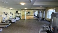 This single-level fitness center offers a comprehensive workout environment designed to accommodate a variety of exercise routines. The space is divided into distinct functional zones, beginning with a cardio area housing treadmills and an elliptical trainer positioned along a mirrored wall that enhances the sense of space throughout the room. Adjacent to this, a large glass-partitioned studio area features a smooth hardwood floor, ideal for group classes, stretching, or floor-based workouts. The strength training zone is well-appointed with a cable resistance machine, free weights, and a workout bench, providing options for both isolated and compound exercises. Rubber flooring throughout the main workout area offers durability and cushioning underfoot. Flush-mounted ceiling lights provide consistent, even illumination across the space. The extensive use of floor-to-ceiling mirrors along multiple walls not only amplifies the perceived size of the room but also allows users to monitor their form during exercise. The overall layout is open and functional, supporting an efficient and well-rounded fitness experience.
