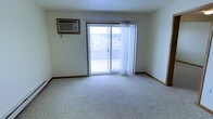 This single-floor residential space offers a clean, functional layout designed for comfortable everyday living. The main living area is finished with wall-to-wall carpet in a neutral tone, providing a warm and inviting foundation throughout the room. Light-colored walls enhance the sense of openness, while wood baseboard trim adds a subtle, classic detail along the perimeter.

A prominent sliding glass door serves as the focal point of the living area, flooding the space with natural light and providing direct access to an outdoor balcony. Sheer white curtains frame the doorway, softening the natural light that pours in and offering a degree of privacy while maintaining brightness. The balcony overlooks neighboring residential buildings, giving the space a connected, community-oriented feel.

Climate control is provided by a wall-mounted air conditioning unit installed on the upper portion of the adjacent wall, ensuring year-round comfort. The textured ceiling adds a traditional finish consistent with the overall aesthetic of the space.

Overall, this space presents a practical and well-lit environment with a straightforward, uncluttered design. Its carpeted flooring, ample natural light, and outdoor balcony access make it a functional and comfortable living space with a welcoming atmosphere.