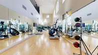 Located at 2615 E Date Palm Paseo in Ontario, California, this fitness center is a standout exercise facility defined by its dramatic double-height ceilings and an open, airy atmosphere. The space is anchored by warm hardwood flooring that runs the full length of the room, lending a polished and professional feel throughout. Floor-to-ceiling mirrors line multiple walls, visually expanding the interior and allowing users to monitor their form during workouts. Large windows and elevated clerestory windows allow abundant natural light to pour in, creating a bright and energizing environment. The cardio equipment section features a well-curated selection of machines, including StairMasters, ellipticals, stationary bikes, and rowing equipment, all from reputable brands such as True Fitness. A dedicated free weights area includes dumbbells, medicine balls, and exercise mats for functional and strength training. An upper-level mezzanine with a railing is visible above, adding architectural depth to the space. Glass-paneled entry doors provide views to the outdoor surroundings. The combination of soaring vertical space, ample natural light, mirrored walls, and comprehensive equipment makes this fitness center a functional and well-designed facility suited for a wide range of exercise routines.