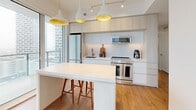 This thoughtfully designed 2-bedroom, 2-bathroom high-rise unit offers a contemporary open-concept layout finished with warm hardwood flooring throughout. The kitchen is a standout feature, showcasing floor-to-ceiling windows that flood the space with natural light and frame sweeping city skyline views. Sleek white cabinetry extends to the ceiling, complemented by stainless steel appliances, a deep undermount sink, and a peninsula with bar seating beneath striking pendant lights with gold interiors — a stylish focal point of the living area. Both bedrooms are well-appointed with large windows offering elevated urban vistas. The primary bedroom benefits from a wall of sliding panel closets and direct access to panoramic views. The second bedroom, currently arranged as a home office, also enjoys generous natural light and outdoor views through full-height glazing. Two bathrooms are finished with large-format grey stone-look tiles. One features a deep soaking tub with an overhead shower, while the other offers a floating vanity with a backlit mirror. A hallway connects all rooms efficiently and includes a dedicated in-unit stacked washer and dryer closet, adding everyday convenience to this well-organized urban residence.